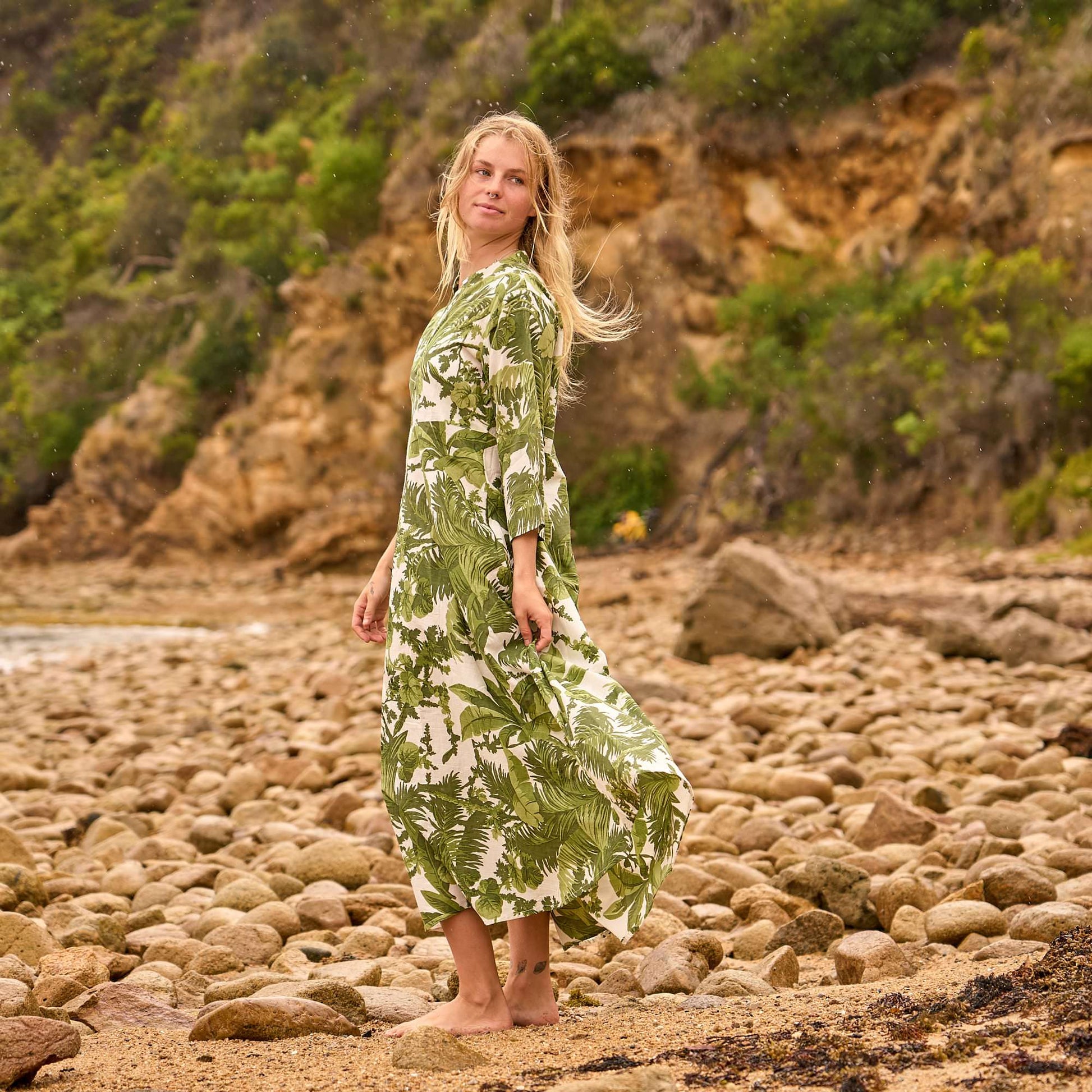 Blonde woman wearing a hand-printed green tropical cotton dress, walking barefoot on the beach in a relaxed maxi silhouette
